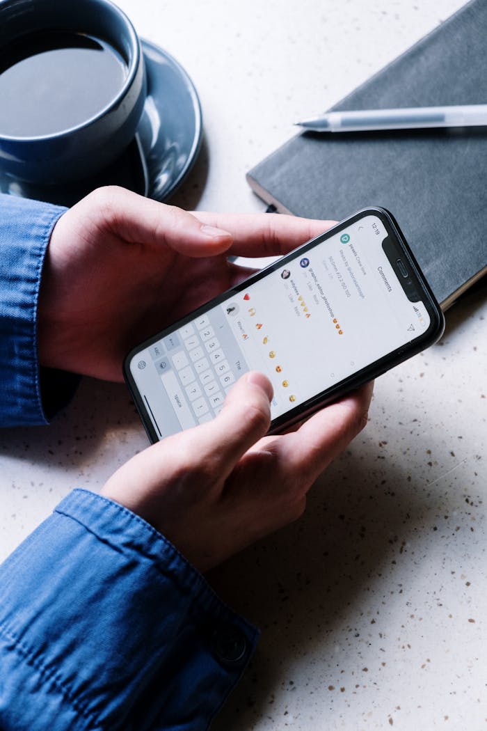 Close-up of hands holding smartphone, using chat app. Coffee and notebook nearby indicate casual setting.